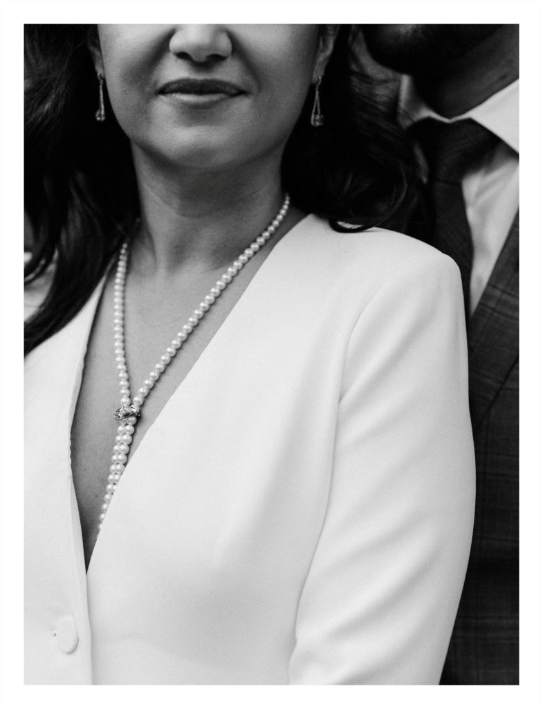 close up image of a bride wearing a white suit with a string of pearls while her husband's suit shirt and tie are behind her