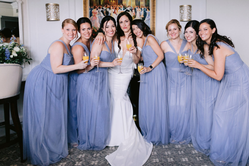 A bride and her seven bridesmaids dressed in lavender dresses pose together inside Le Chateau before the wedding begins