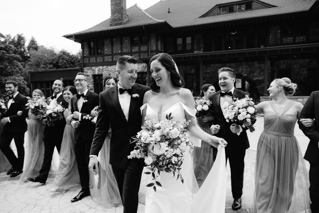 An extremely fun and large wedding party walks along laughing together in front of the elegant Le Chateau wedding venue in South Salem New York