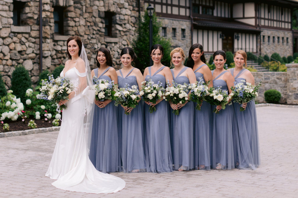 An elegant bride and her large group of bridesmaids pose in front of the elegant Le Chateau wedding venue in South Salem New York