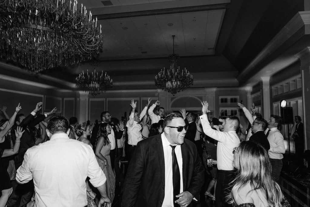 Giant party on the dance floor of the ballroom at LeChateau in South Salem New York. A man wears his sunglasses front and center and signs with the music while dancing.