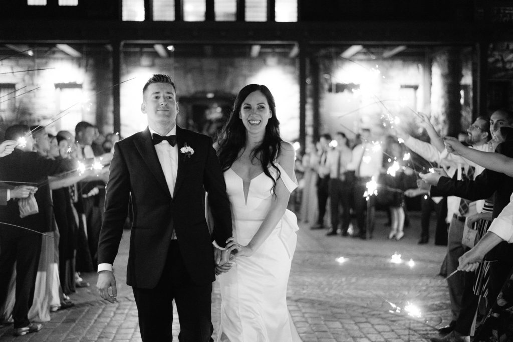 Bride and groom smile as they leave their crazy fun wedding at Le Chateau in South Salem New York to a crowd of sparkler-holding happy guests