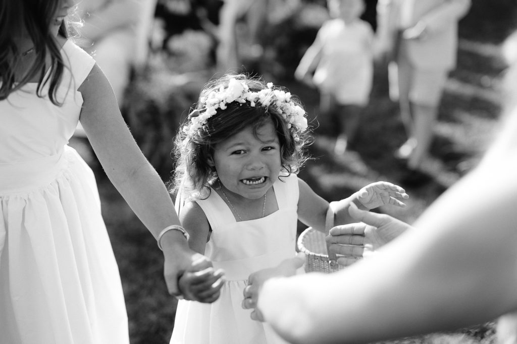 funny photo of a little flower girl with brown curls crying as she walks down the aisle during a cape cod wedding ceremony