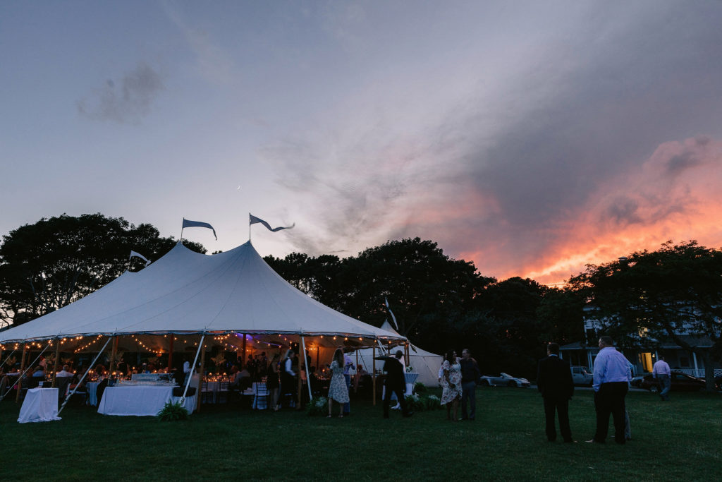 wide angle photo of a wedding reception being held in a tent on cape cod with a beautiful sunset happening behind it