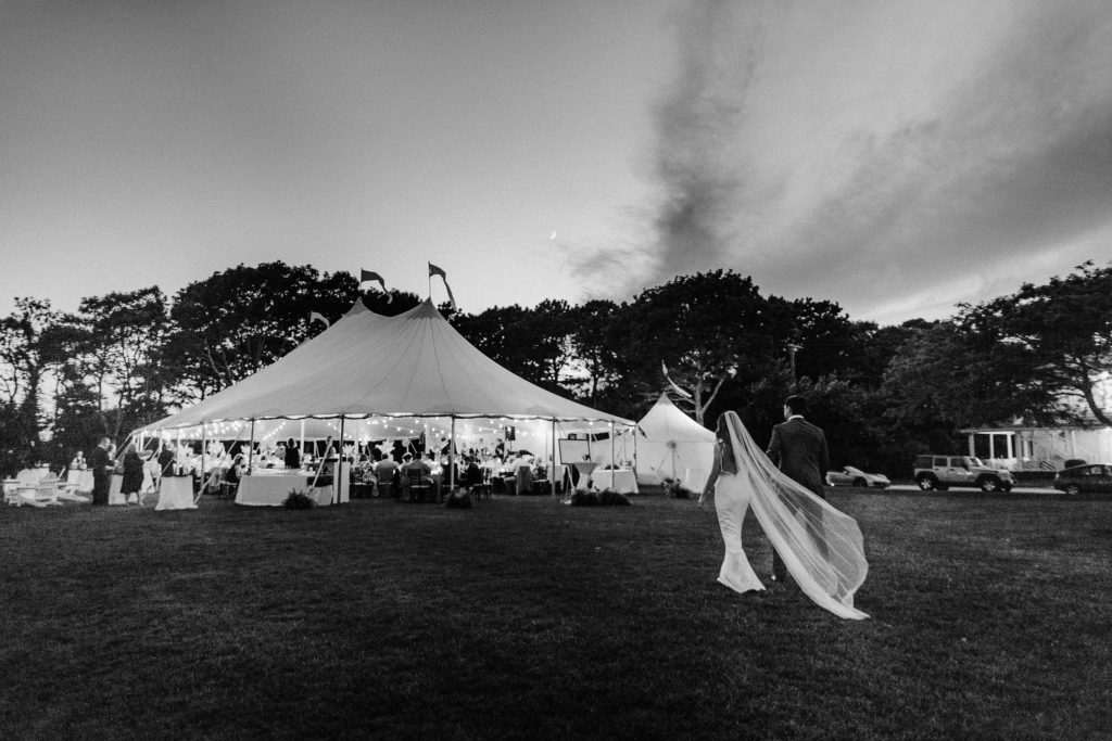 black and white image of bride and groom walking toward their reception tent as the sun sets in cape cod