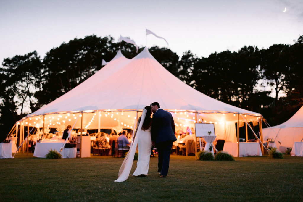 bride and groom walk hand in hand toward their reception tent which is lit up in front of them on the shores of cape cod