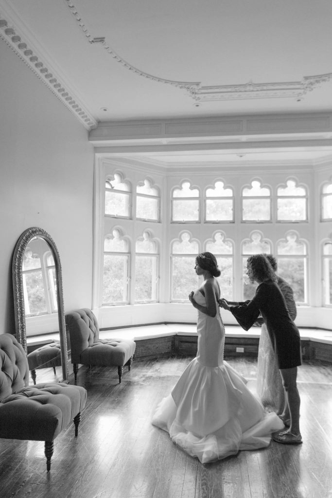 Bride is helped into her dress by her mom and her bridesmaids in a huge room flanked by windows in Castle Gould at the Sands Point Preserve in New York