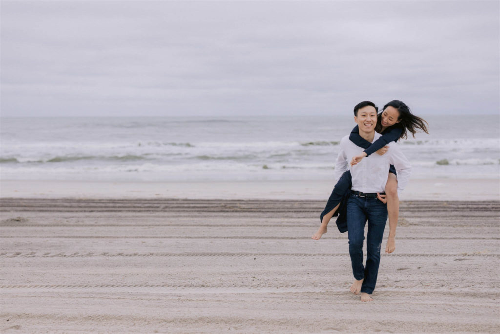 Smiling Asian man piggybacks his smiling Asian fiancee as she walks barefoot along the beaches of Fire Island in New York State