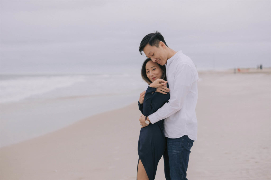 Asian man hugs his Asian fiancee from behind and rests his cheek on the top of her head while they both smile and look at the camera with the ocean behind them at Fire Island New York