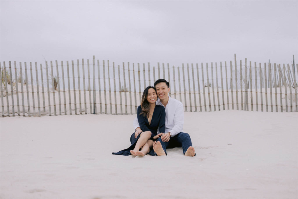 Asian man and woman sit together in the sand, smiling, with a quintessential beach fence behind them on Fire Island in New York State