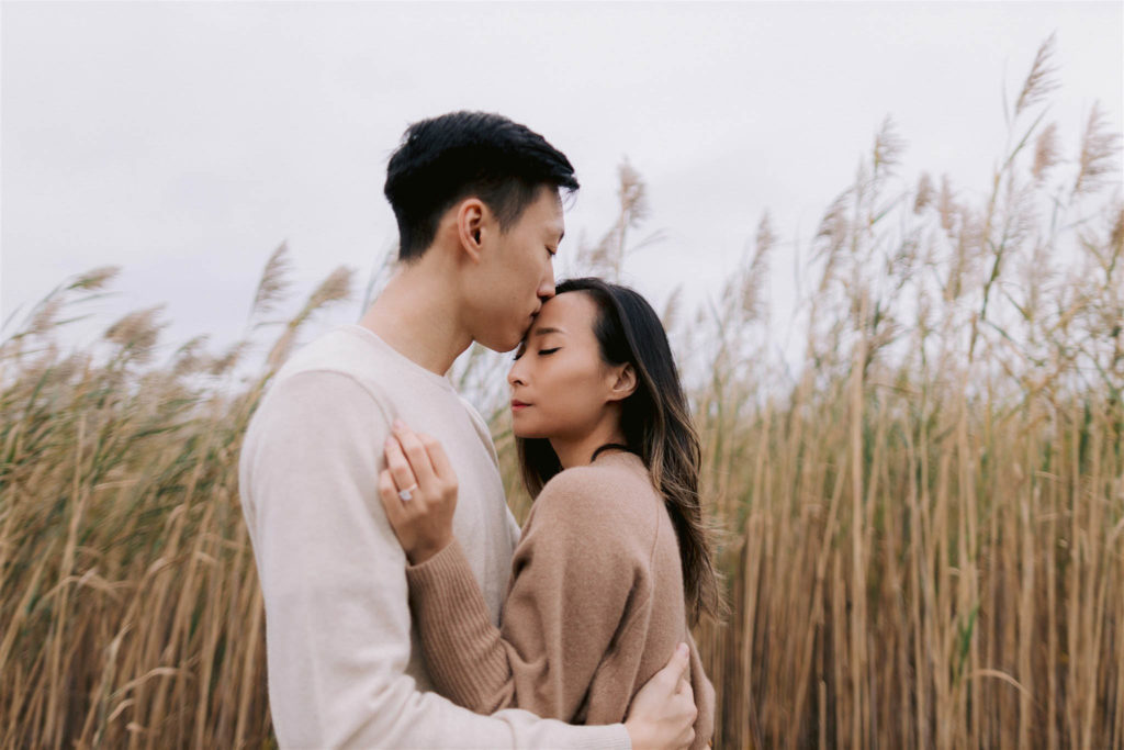 Asian man in his 20's kisses his fiancee's forehead as she lays a hand on his chest with tall reeds behind them on Fire Island in Long Island New York