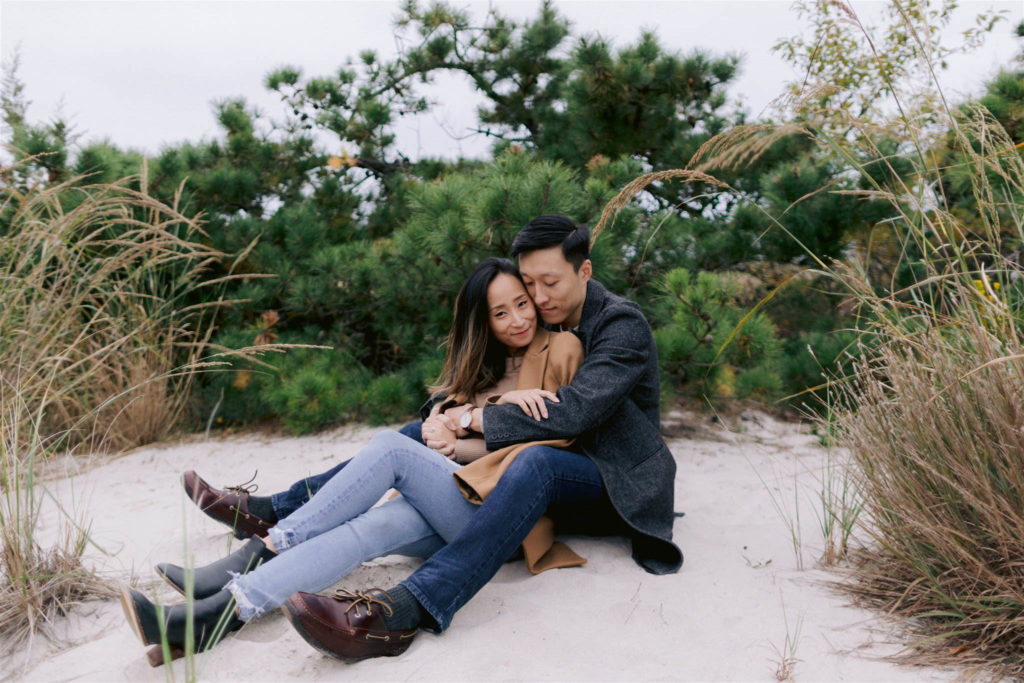 And Asian man and woman dressed chic casual sit on the beach at Fire Island New York, snuggled into each other. Image by Jenny Fu Studio