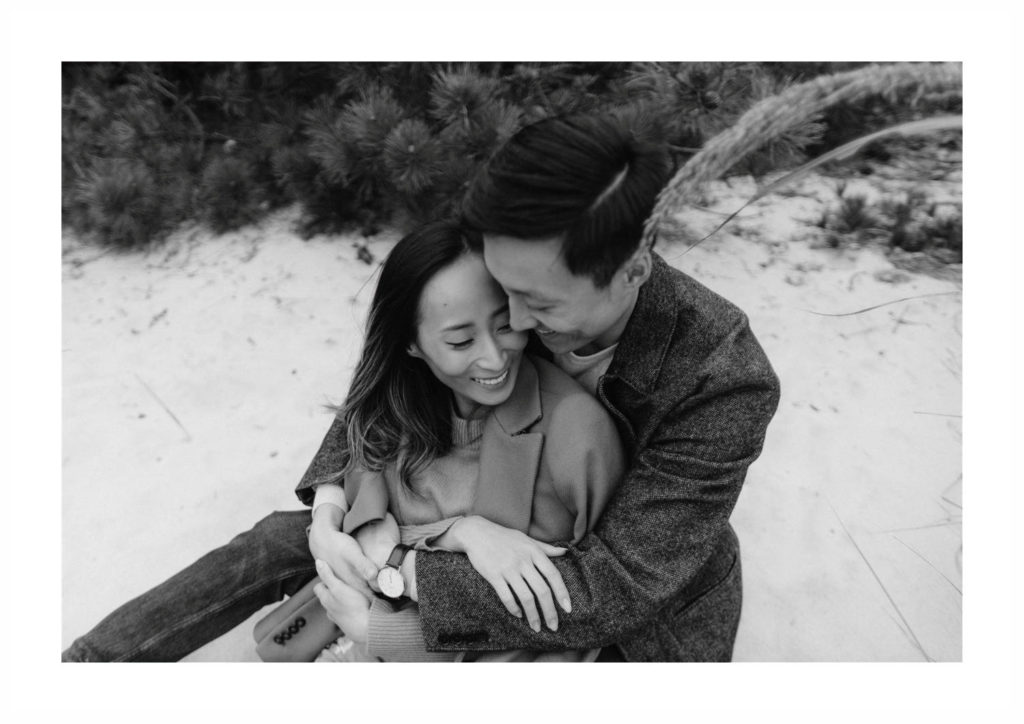 Asian man and woman celebrate their engagement with snuggles and smiles as they sit on the beach at Fire Island in New York.