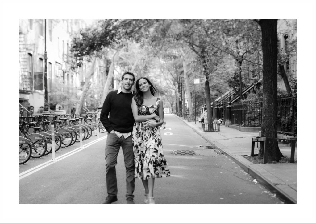Young couple walks down a park-lined street of New York City smiling with his arm around her.
