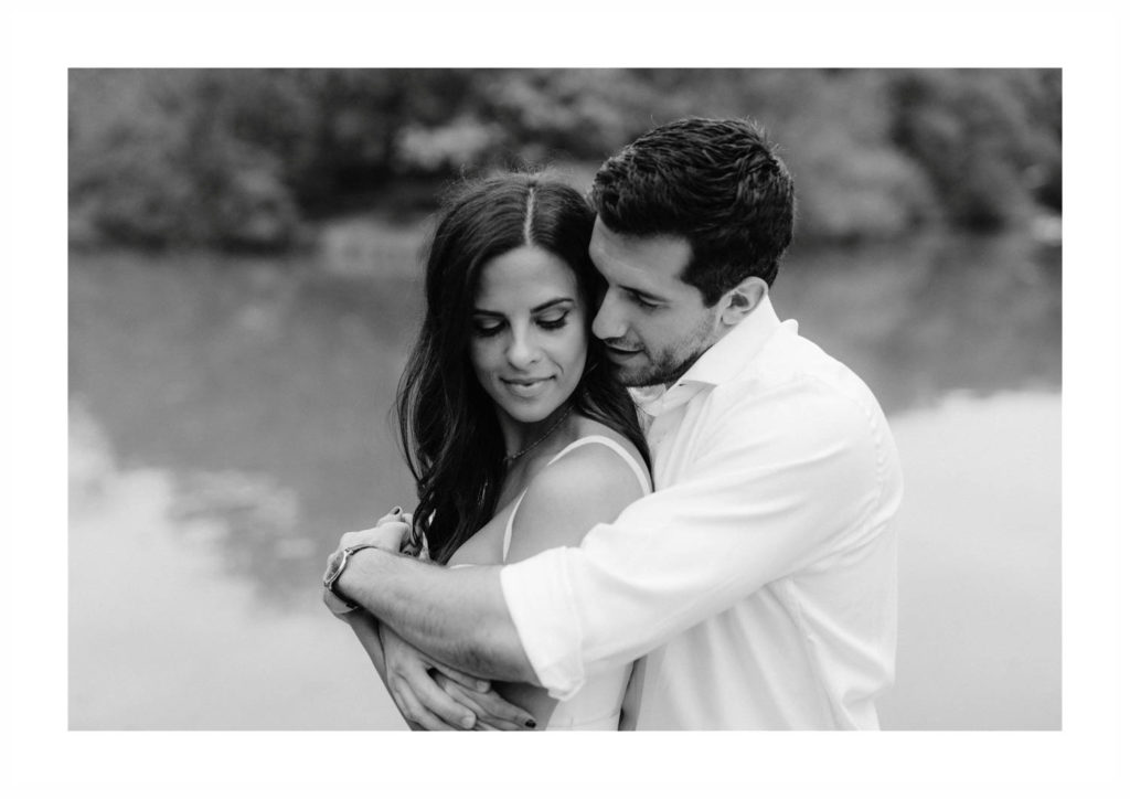 Young man in a white shirt wraps his arms around his fiancee and puts his nose against her cheek with a pond behind them in New York's Central Park