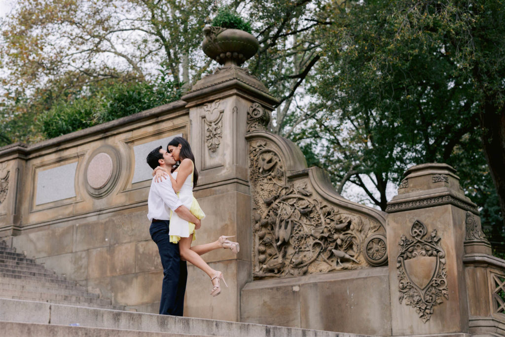 Man scoops his arms under his fiancee's bottom and lifts her up to kiss him near in front of the gates of Central park in NYC