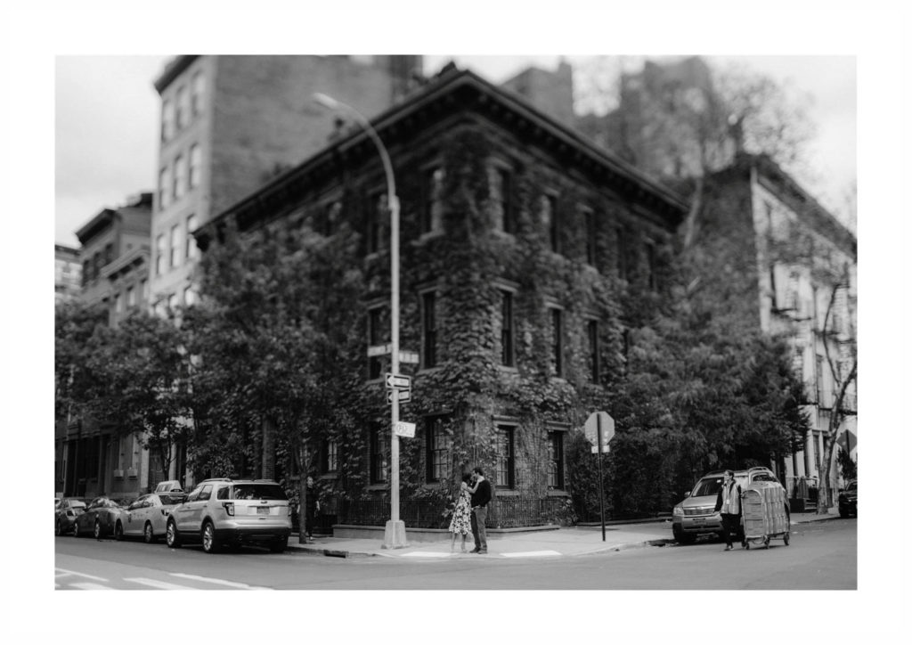 Engaged New York City couple stands before a famous NYC brownstone staring at each other and smiling, black and white version