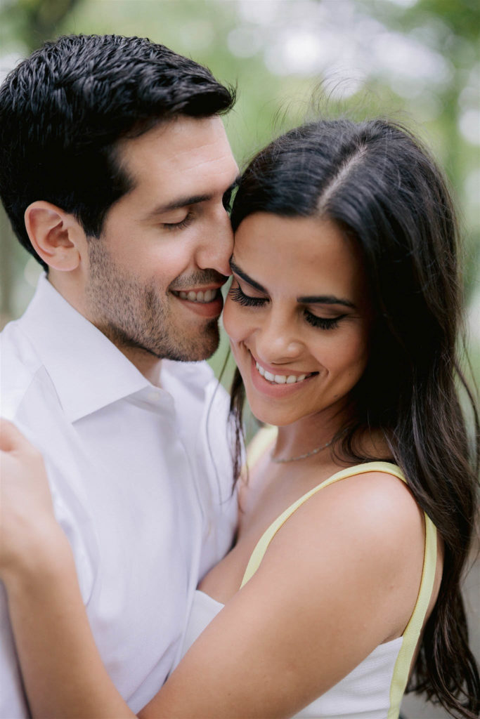 Close up shot of a newly engaged young couple in New york City. Both have their eyes closed, he has his nose placed gently against the side of her head and they are smiling happily.