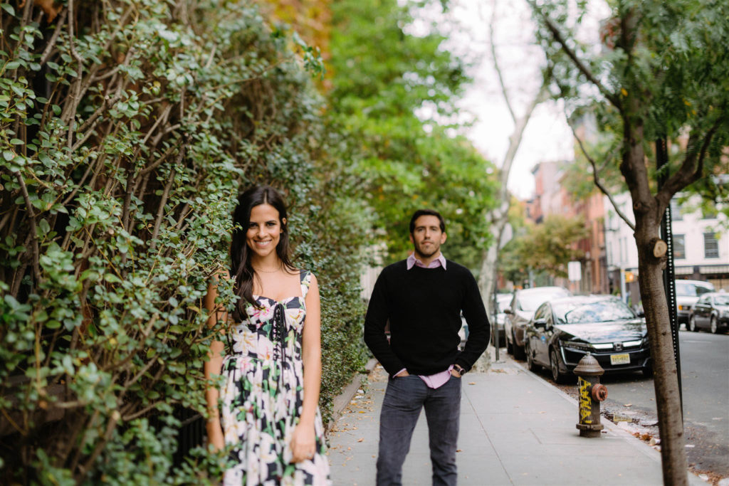 Woman wearing a cute floral print dress leans against a hedge smiling while her fiance stands a few feet behind her with his hands in his jean pockets. Both are smiling at the photographer, Jenny Fu as they pose for engagement photos in the West Village of New York City