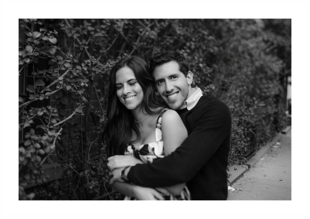 Young, twenty-something man hugs his gorgeous, dark-haired fiancee and looks at the camera smiling against a hedge in New York City's West Village
