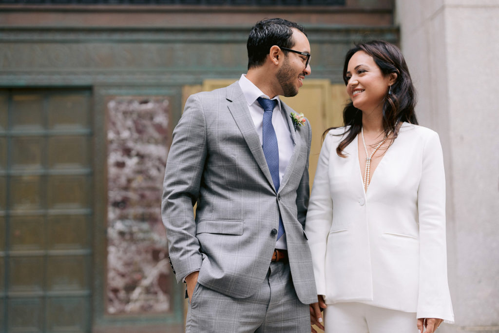 A couple who just eloped stand on the steps of New York City Hall smiling at each other