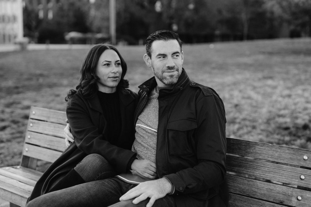 good looking couple in their late 20's sit on a park bench in a park in Brooklyn Heights and look off into the distance.