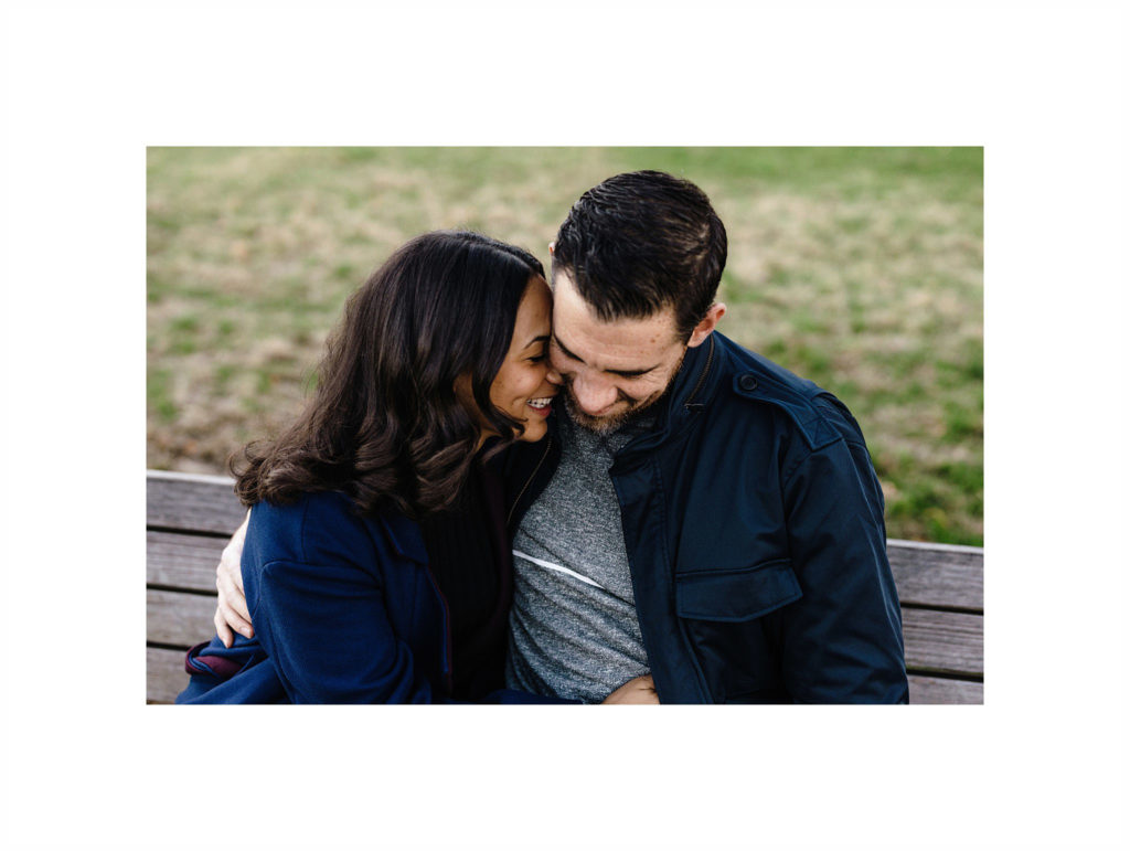 Cute mixed race couple snuggles into each other on a park bench in Brooklyn Heights and giggles. Image by Jenny Fu