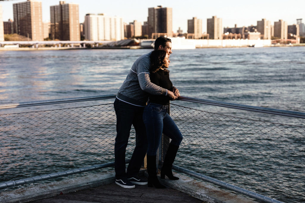 Attractive mixed race couple celebrates their love with an engagement session and snuggles up together, leaving against a fence overlooking the river in Brooklyn