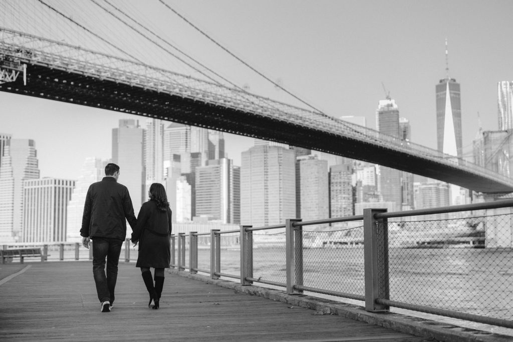 A happily engaged couple celebrate their love by walking hand in hand under the Brooklyn Bridge