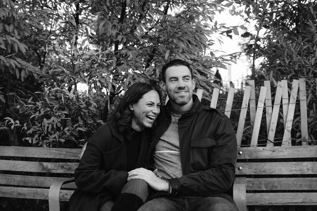 Couple sitting on a park bench in Brooklyn Heights during an editorial engagement session looks off into the distance, the woman laughing hysterically
