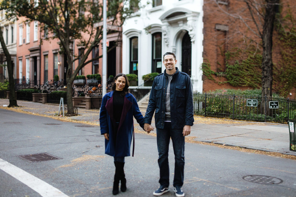 An attractive mixed race couple stand holding hands in front of a Brownstone with a white facade in Brooklyn Heights