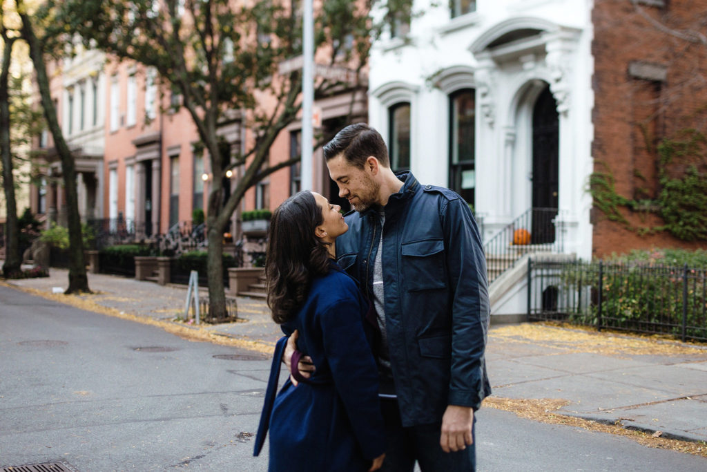 A gorgeous man and woman stand on the street in Brooklyn Heights, snuggled together closely and staring into each other's eyes
