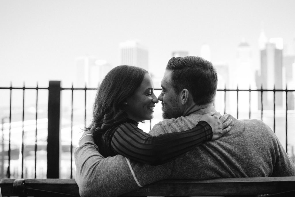 A newly engaged couple snuggles up together on a park bench in brooklyn heights with New York City behind them across the river