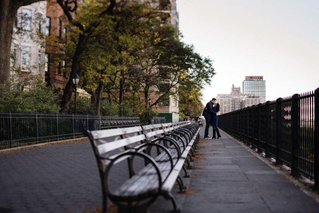 Romantic shot of a row of park benches in the foreground leading to a couple snuggling at the end of the benches in the background in Brooklyn Heights. Image by Jenny Fu Studio