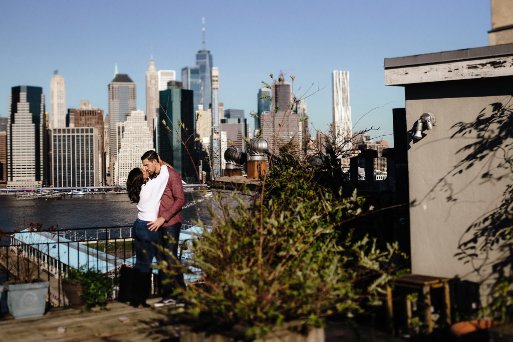 Urban shot of an engaged couple posing —her arms wrapped around his neck—with NYC in the background