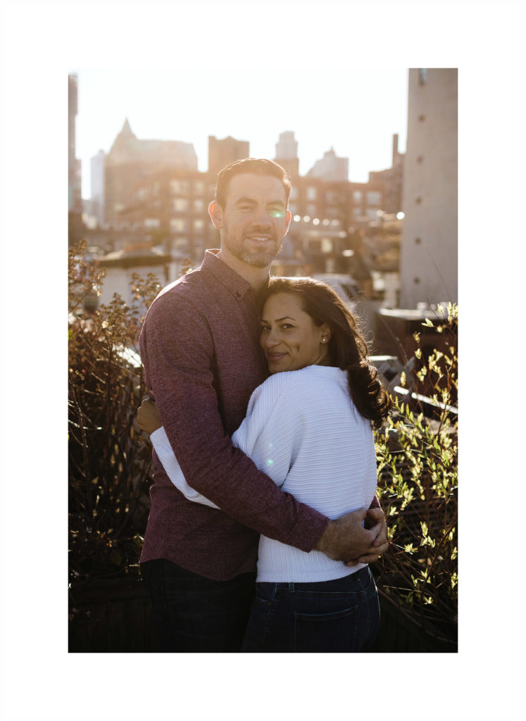 Cute mixed race engaged couple snuggles into each other in Brooklyn Heights, her face pressed against his chest, looking at the camera