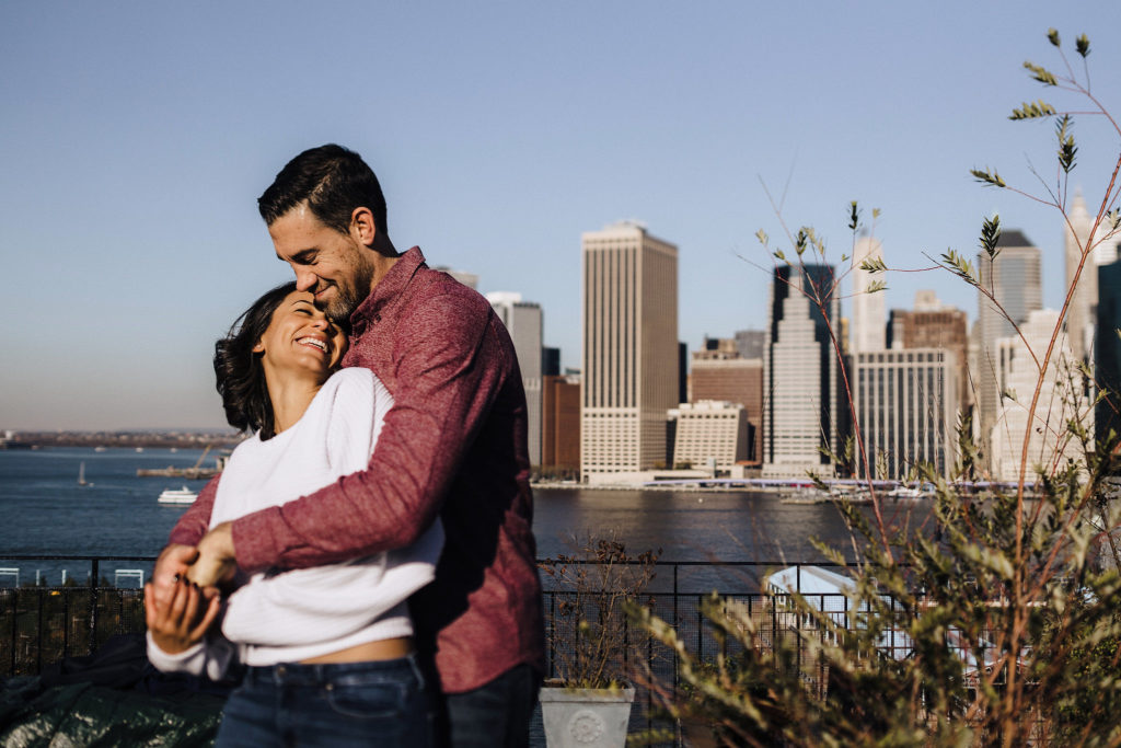 Cute couple snuggles into each other smiling with NYC across the river behind them under a gorgeous blue sky day