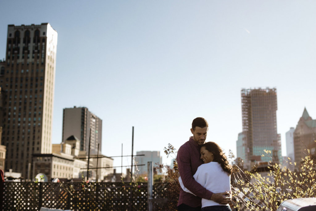 A gorgeous mixed race woman snuggled into her tall fiance's chest with her eyes closed and the New York City Skyline behind them. Jenny Fu Photography