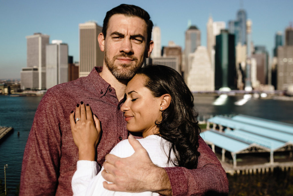 A tall white man wraps his arm tightly around his mixed race fiancee as she leans against his chest with her eyes closed, smiling. jenny fu studio