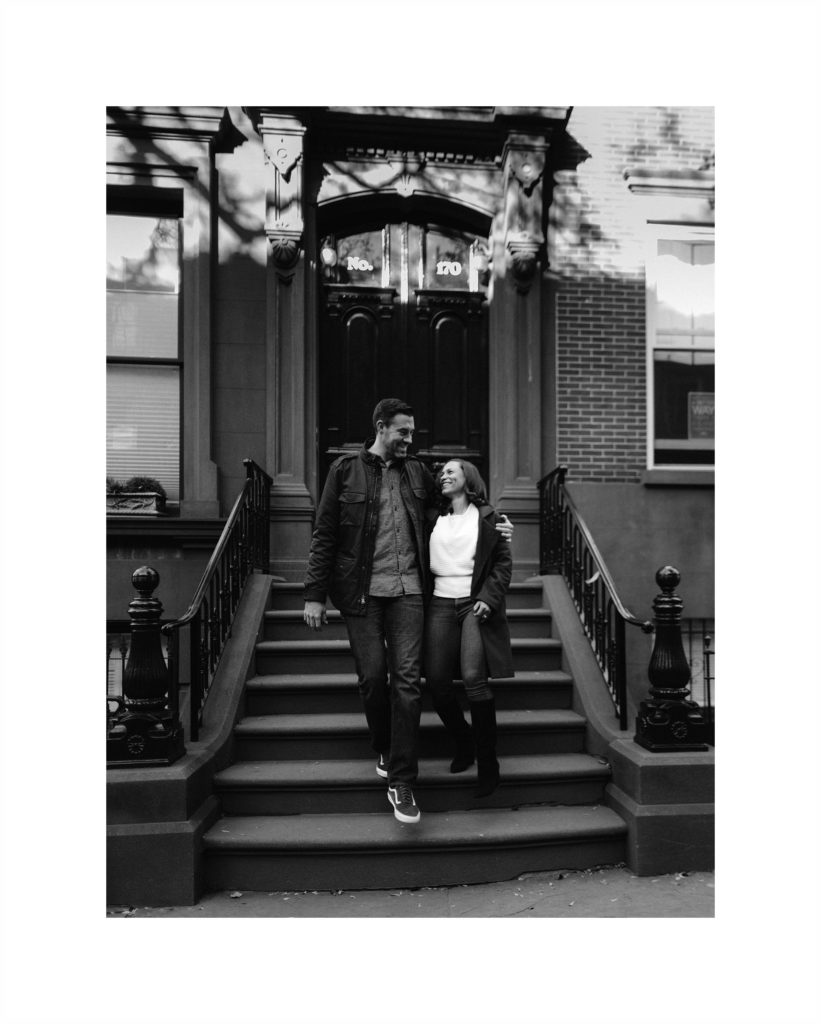 Newly engaged couple stands on the steps of a Brownstone in Brooklyn Heights, looking at each other with adoration.