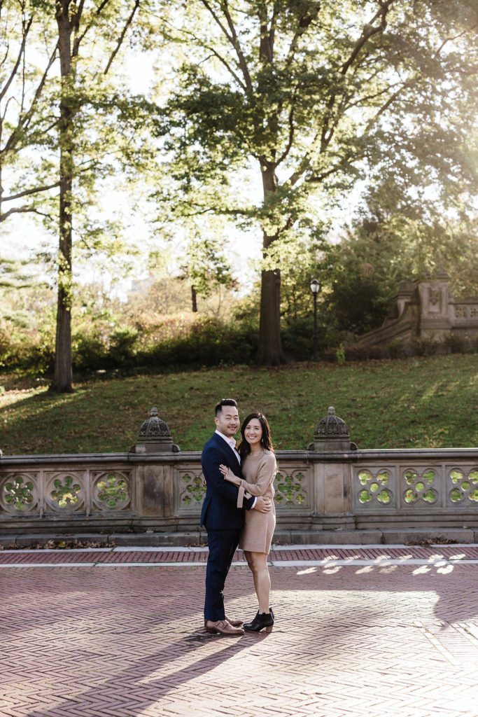 good-looking engaged asian couple embraces each other on a cement walkway with central park, new york behind them. images by jenny fu