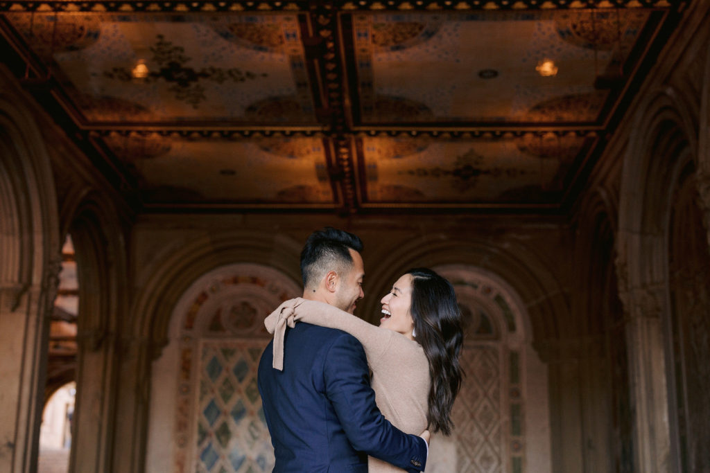 Laughing, newly engaged asian couple hug each other in front of a set of ornate and decorative windows in the west village of new york city