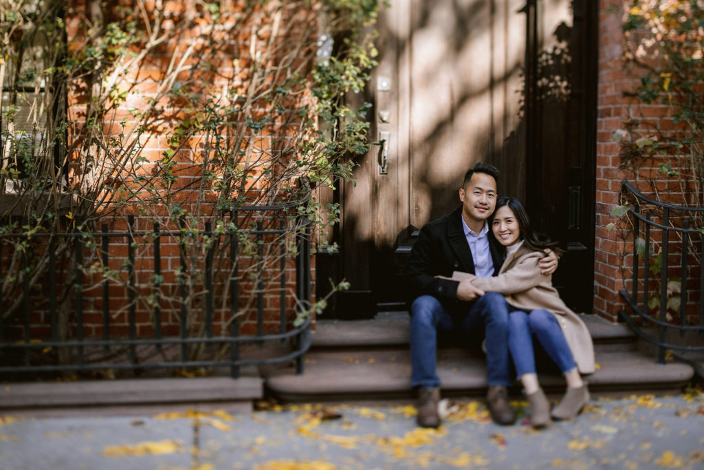 an engaged asian couple sits on a step in a doorway of the west village, hugging each other and looking at the camera, smiling