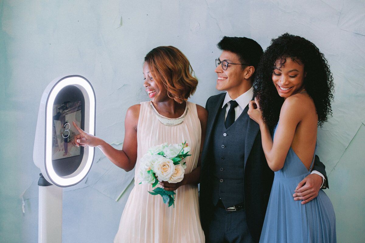 guests at a wedding hug each other in front of a digital photo booth and get ready for the photo to be taken