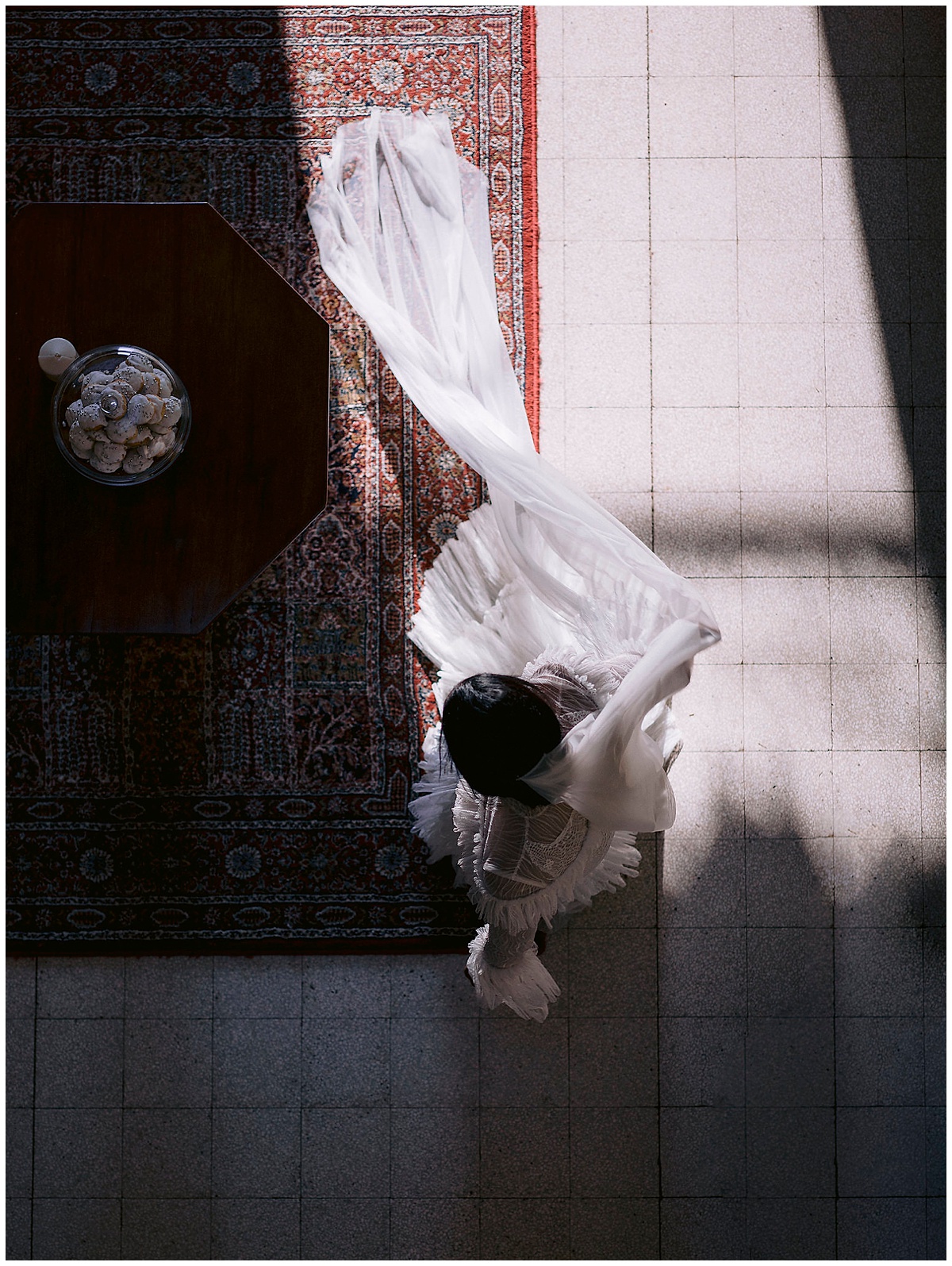 bride stands in a strip of sunlight on a persian rug with her cathedral veil trailing beside her. Editorial and photojournalistic photography. Image by jenny Fu