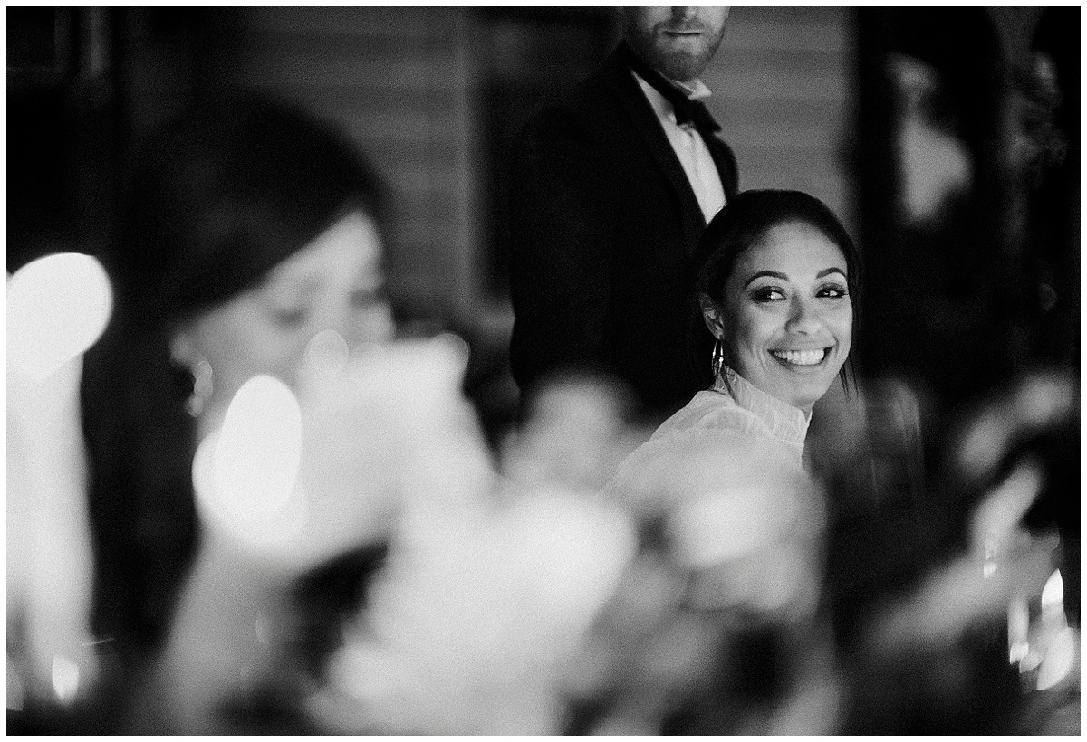 Bride smiles down the table at her blurred out bridesmaid in the evening over candlelight. Editorial photography.