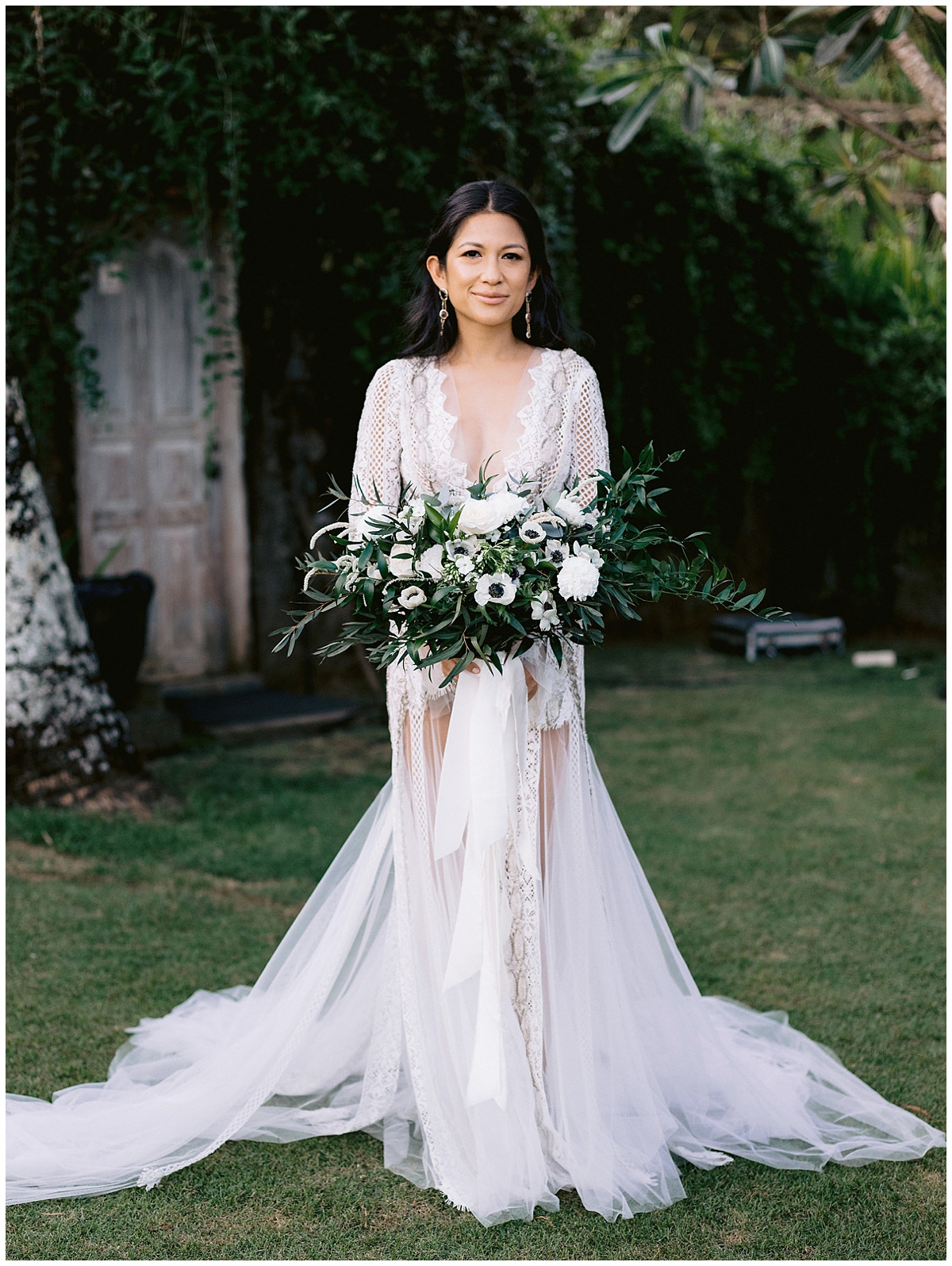 Bride holding a large bouquet of mixed white flowers and wearing a lacey, sheer gown that shows her legs from mid-thigh with a beautiful green space behind her. Editorial images taken by Jenny Fu