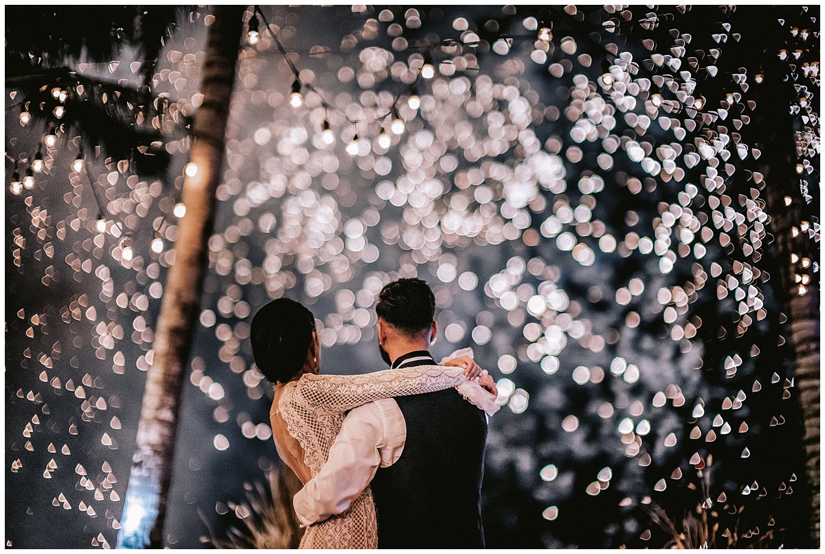 Bride wraps her arms around her groom's shoulders while they both watch fireworks exploding above them in Bali. Editorial image by Jenny Fu studio