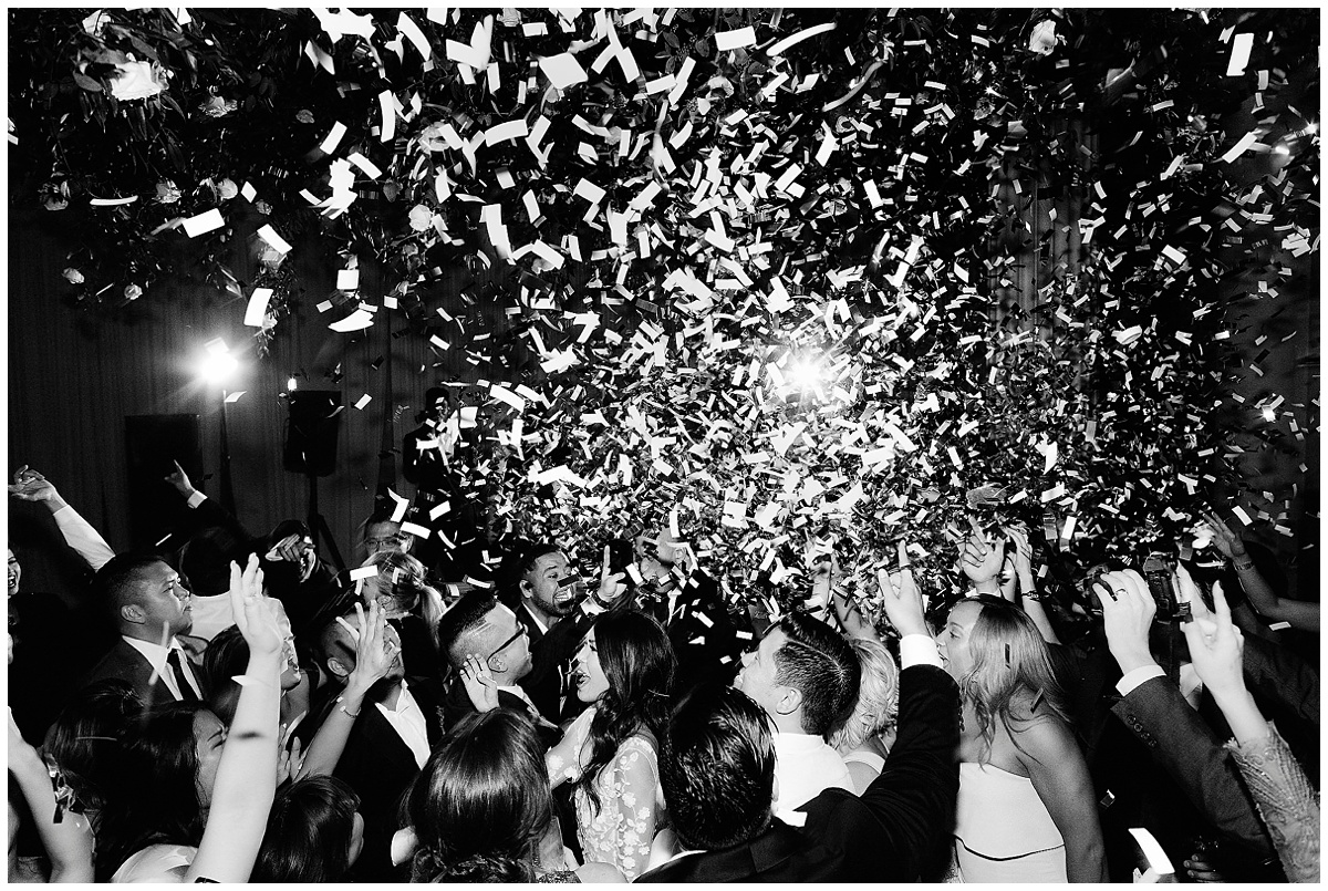 wedding guests sing and dance underneath a pile of flying paper confetti