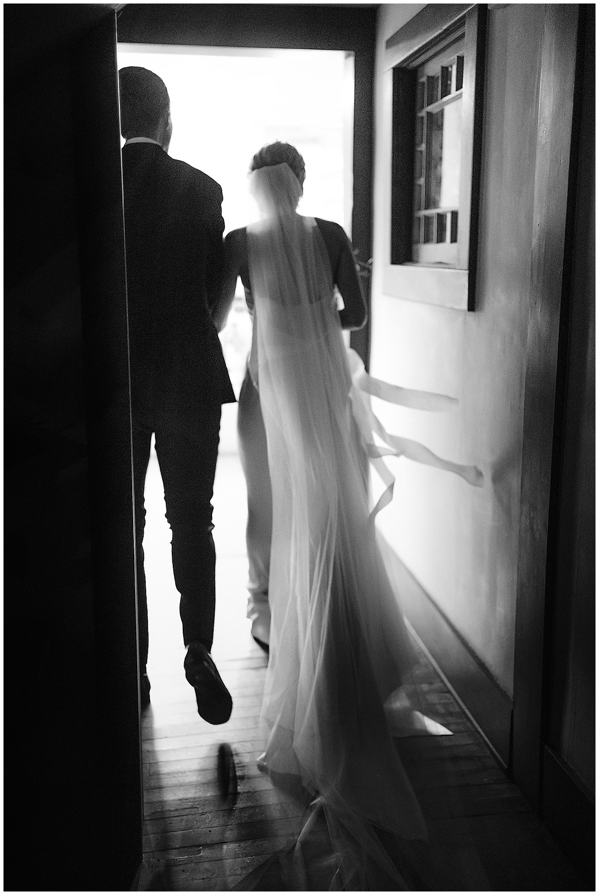 a bride and groom in silhouette as they walk down a hallway into a well-lit reception hall with the bride's cathedral veil trailing behind them